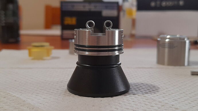 The image shows a metal RDA (Rebuildable Dripping Atomizer) part for an electronic cigarette or vape, under assembly, showing the build posts and O-rings. (Subtitled by AI)