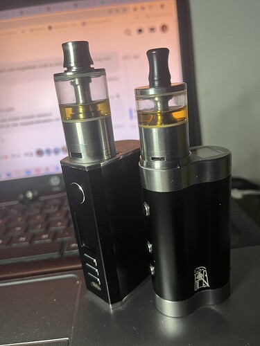 Two black electronic smoking devices (vape) stand on a table with a laptop screen in the background. (Labeled by AI)