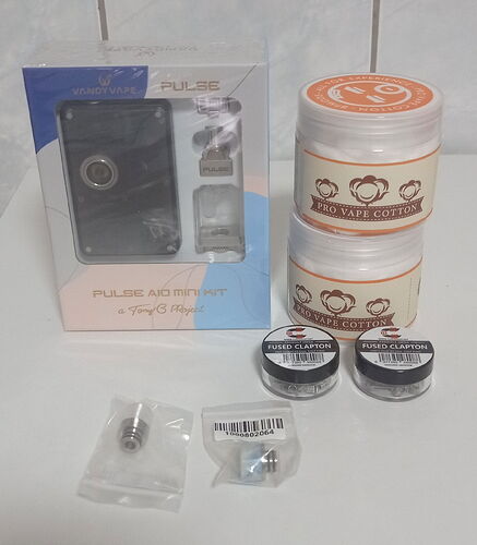 Egy asztalon egy új Vandy Vape Pulse AIO Mini Kit doboza, két tégely Pro Vape Cotton, két kis tartó Fused Clapton tekercs és további gőzöléshez való alkatrészek láthatók. (Feliratozva AI-val)