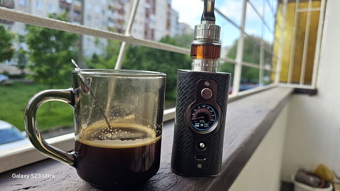 Az erkély korlátján egy fekete kávés bögre és egy digitális kijelzős vape eszköz áll, háttérben társasházak és zöld fák láthatók. (Feliratozva AI-val)