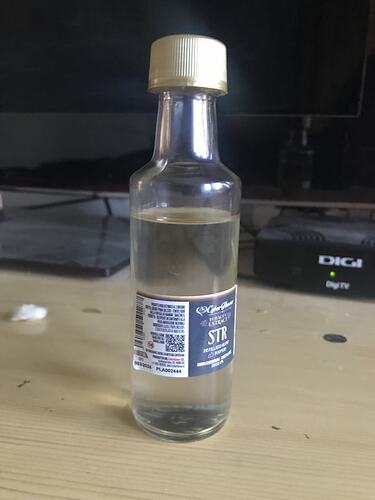 A small bottle of clear liquid on a table, in the background, a TV and a DIGI set-top box are visible. (Subtitled with AI)