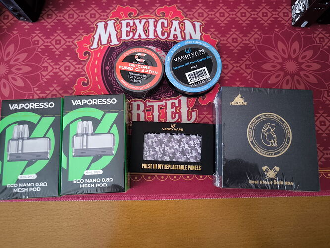 Az asztalon két Vaporesso Eco Nano 0.6 Ohm Mesh Pod, két Vaporesso cserélhető panel, két kis tartály és egy doboz cigaretta látható egy piros "Mexican Cartel" feliratú alapon. (Feliratozva AI-val)