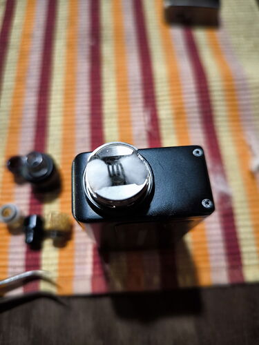 Egy fekete gép vagy eszköz tetején egy fényes, csillogó fémgomb vagy rész látható, amelyen egy villa tükröződik, egy csíkos, több színű textil háttér előtt. (Feliratozva AI-val)