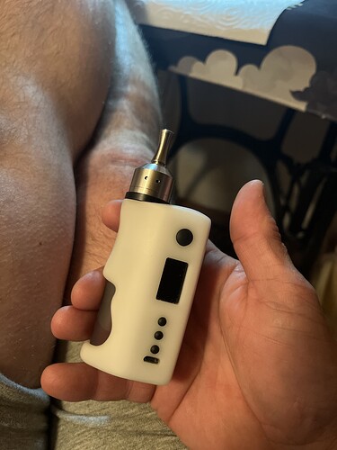 Egy kéz egy fehér színű elektromos cigaretta eszközt tart, miközben a háttérben egy személy lába látható. (Feliratozva AI-val)