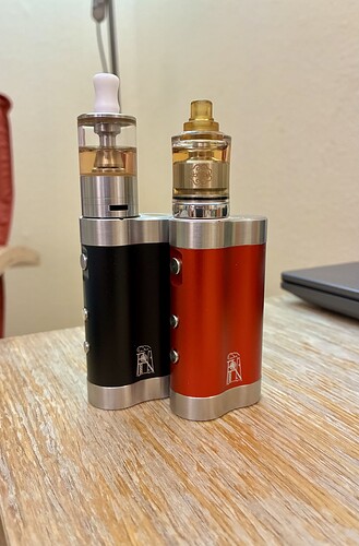 Két különböző színű elektromos cigaretta áll egy fából készült asztalon, háttérben homályosan látható egy laptop és egy piros tárgy. (Feliratozva AI-val)