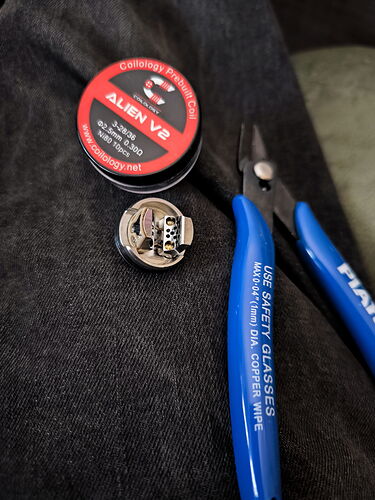 A Coilology Alien V2 wire coil, a vape coil, and a blue pair of pliers lie on a dark fabric. (AI-captioned)