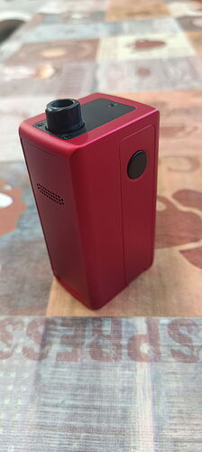 The image shows a red, box-shaped electronic cigarette (vape) on a patterned tablecloth. (Subtitled by AI)