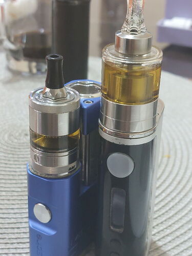 Deux vaporisateurs électriques (e-cigarettes) de tailles et de couleurs différentes sont visibles côte à côte. (Légendé par IA)