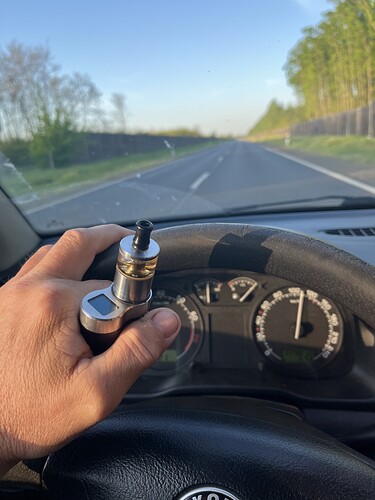 Une main tenant une cigarette électronique tout en conduisant derrière le volant sur une route droite et vide au coucher du soleil. (Sous-titré par IA)