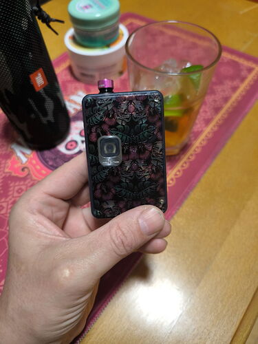 A small electronic device with a patterned design held in a hand is visible, with an empty glass with ice and several other objects on the table in the background. (Subtitled with AI)