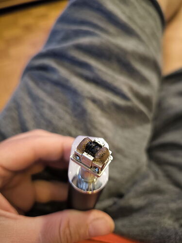 Egy kéz egy elektromos cigaretta felülről látható felületét tartja, amelyben megolvadt vagy leégett szénszálak láthatók. (Feliratozva AI-val)