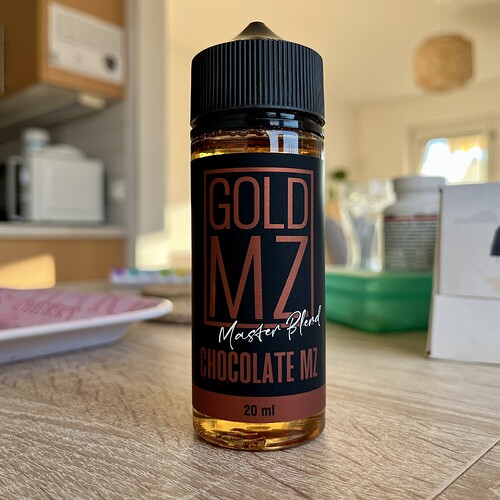 A 20 ml plastic bottle containing liquid labelled "GOLD MZ Master Blend Chocolate Milk" on a table, in a kitchen environment. (Labeled by AI)
