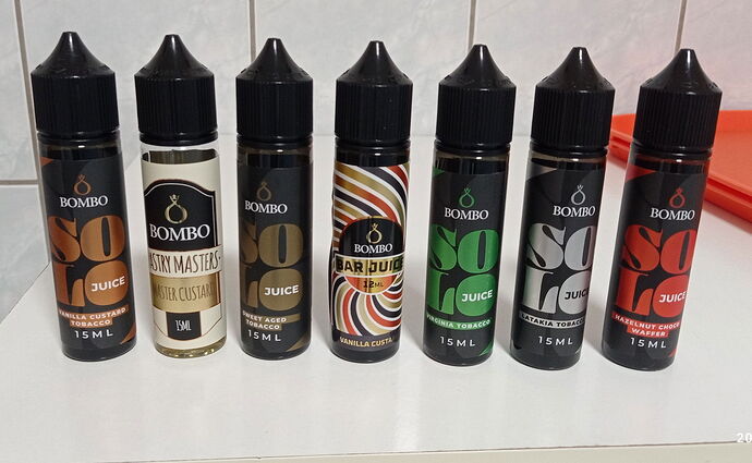 A képen hét különböző ízesítésű Bombo márkájú e-liquid palack látható egymás mellett egy fehér felületen. (Feliratozva AI-val)