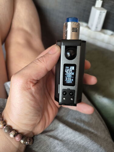 Egy kéz egy elektronikus vape eszközt tart, amelynek kijelzőjén különböző beállítási értékek láthatók, miközben egy láb pihen a háttérben. (Feliratozva AI-val)