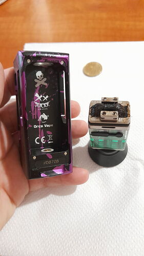 A hand holding a purple and black vape mod with an open battery compartment, next to a dirty atomizer on a white surface. (Subtitled by AI)