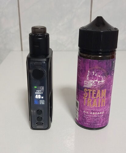 A képen egy fekete elektromos cigaretta és mellette egy nagyfekete kupakkal ellátott, lila címkés Steam Train e-liquid palack látható egy fehér asztalon. (Feliratozva AI-val)