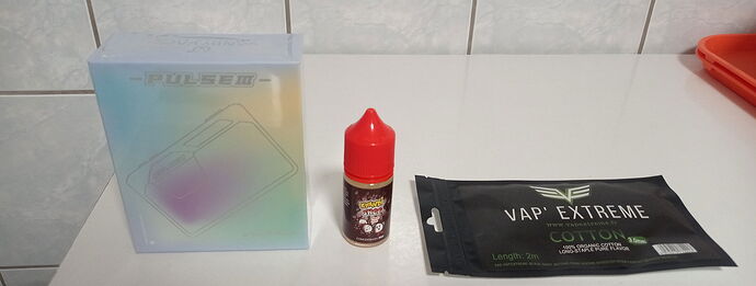 Az asztalon egy Pulse BF RDA doboz, egy piros kupakos folyadék üveg és egy Vapor Extreme gyapot csomag látható. (Feliratozva AI-val)