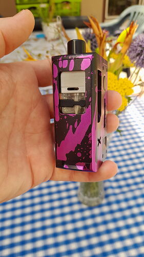 A hand holds a purple and black patterned e-cigarette over a blue-white checkered tablecloth, with blurred flowers in the background. (Labeled by AI)