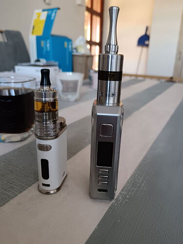 Az asztalon két különböző méretű és kialakítású elektromos cigaretta van elhelyezve, mögöttük egy szoba belső rész látható. (Feliratozva AI-val)