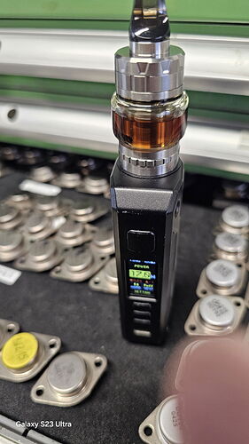 Un appareil de vapotage est visible sur la photo, qui est un mod au corps noir, avec un tank coloré sur le dessus, et diverses pièces métalliques en arrière-plan. (Sous-titré par IA)