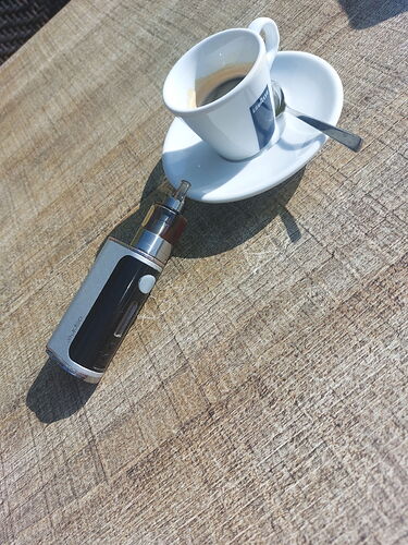 Une tasse d'espresso sur la table, avec une soucoupe avec une cuillère et une cigarette électronique noir et argent visible. (Légendé par IA)