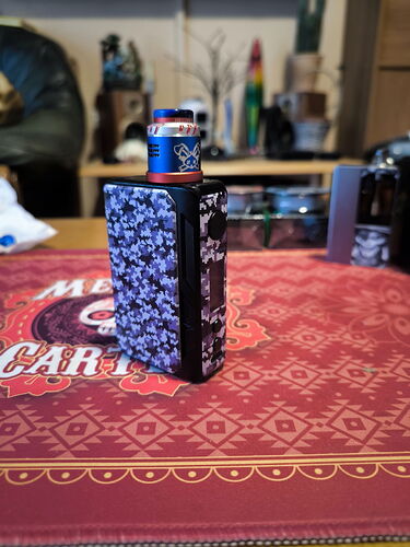 A képen egy mintás vape eszköz látható egy piros asztalterítőn, háttérben homályosan különféle tárgyakkal. (Feliratozva AI-val)