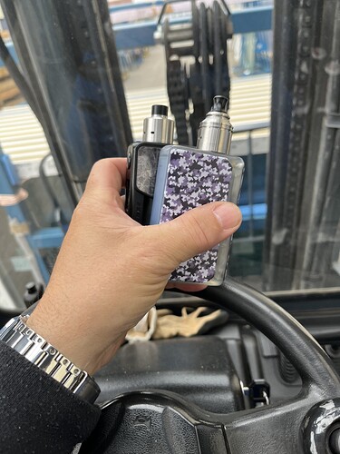 A képen egy kéz látható, amely két elektromos cigarettát tart, miközben egy kormánykerék mellett áll. (Feliratozva AI-val)