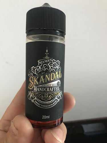 Une main tient une bouteille de liquide de 20 ml, de conception noire, à étiquette ornée, de la marque SKANDAL, faite à la main, saveur tabac sucré. (Légendé par IA)