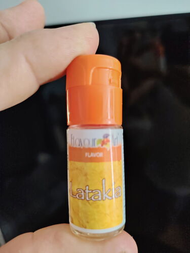 A hand holding a bottle containing a liquid called Latakia, with an orange cap, which tastes like smoked tobacco, against a black background. (Auto-captioned)