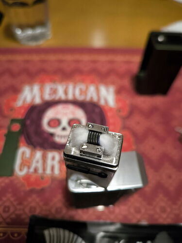 A képen egy fémes gyújtó látható, amely a háttérben egy mexikói tematikájú terítőn helyezkedik el. (Feliratozva AI-val)