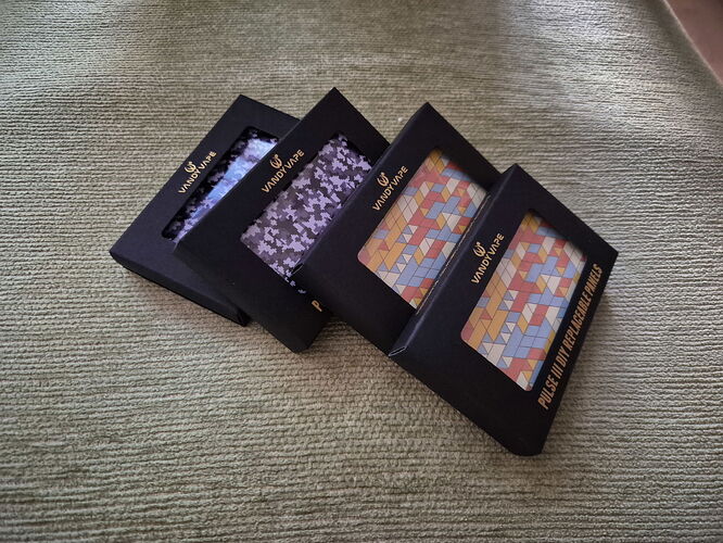 Négy, fekete dobozban lévő színes mintás anyag vagy csempe van elhelyezve egymás mögött egy szőnyegen. (Feliratozva AI-val)