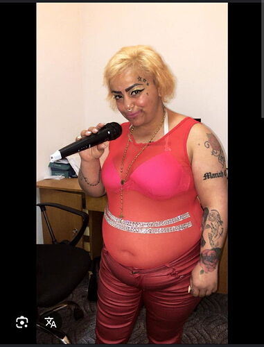 Egy tetoválásokkal díszített személy piros felsőben és nadrágban, mikrofont tartva áll egy szoba belsejében. (Feliratozva AI-val)