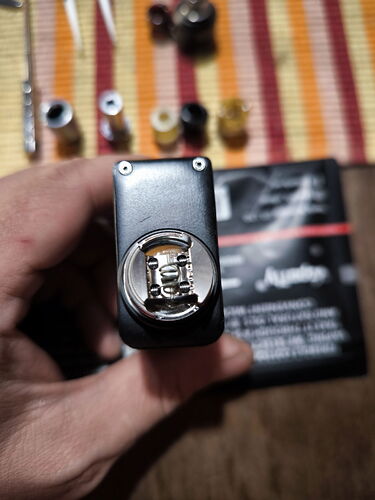 A kép egy kézben tartott, felülről fotózott elektromos cigaretta fűtőtekercsének szerelését mutatja, miközben a háttérben különböző alkatrészek és egy doboz látható. (Feliratozva AI-val)