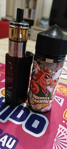 Egy fekete e-cigaretta áll egy Unicorn's Cannoli feliratos e-liquid flakon mellett, mindkettő egy színes, mintás alátéten fekszik. (Feliratozva AI-val)