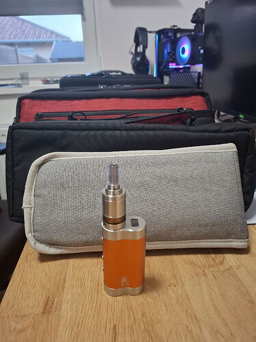 Az asztalon egy narancssárga színű e-cigaretta áll, mögötte pedig három tok különböző méretekben és színekben. (Feliratozva AI-val)