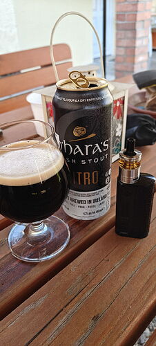 Egy faasztalon egy üvegpohár, egy nyitott O'Hara's Irish Stout Nitro sörösdoboz és egy fekete vape készülék látható. (Feliratozva AI-val)