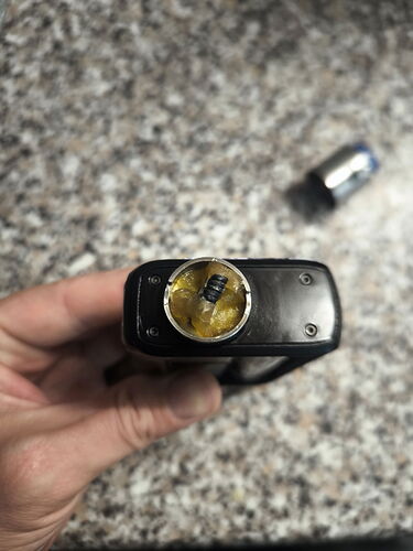 A black vape device held in a hand, showing its coil and a thick, yellow substance collected in a spoon. (AI-generated caption)
