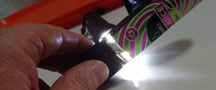 A képen egy kézi lámpa látható, amelynek fényét egy kéz tartja, miközben egy színes, ízesített italcsomag mellett helyezkedik el. (Feliratozva AI-val)