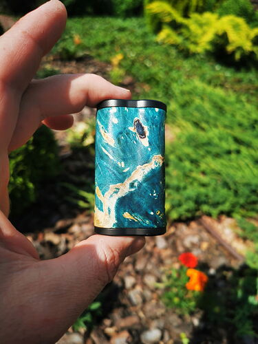 Une main tenant un objet de petite taille à motif bleu et marron entre deux doigts, avec des plantes vertes et des fleurs en arrière-plan. (Légendé par IA)