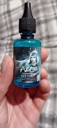 Une main tenant un flacon compte-gouttes rempli d'un liquide bleu, dont l'étiquette montre un personnage féminin aux cheveux blancs et couronné, avec l'inscription : "A&L Ultimate Shiva - Sweet Edition". (Traduit automatiquement par IA)