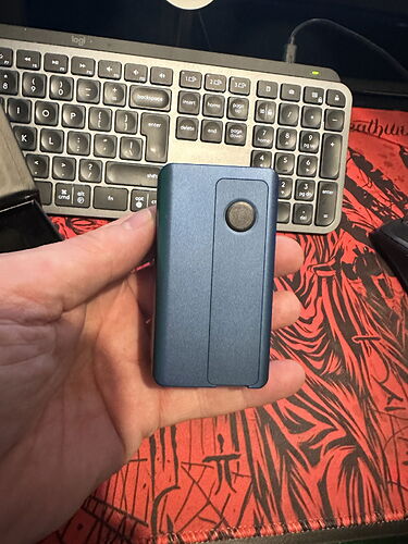 Egy kéz tart egy kék, kis méretű elektronikus eszközt egy billentyűzet fölött egy piros mintás egéralátéten. (Feliratozva AI-val)