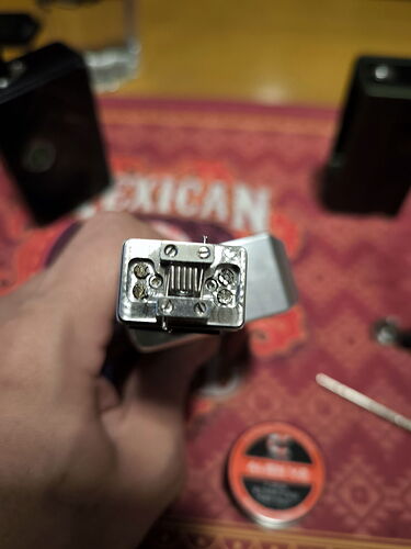 Az képen egy elektromos cigarettához tartozó fém alkatrészt láthatunk, amelyet egy kéz tart, míg a háttérben eszközök és egy "Mexican" feliratú anyag található. (Feliratozva AI-val)