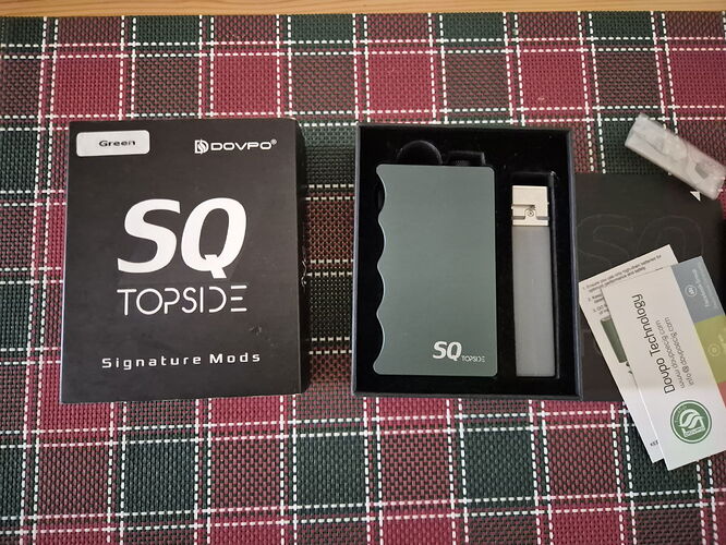 Un kit e-cigarette Dovpo SQ Topside Signature Mods est visible avec sa boîte, l'appareil et ses accessoires sur une nappe à carreaux. (Légendé par IA)