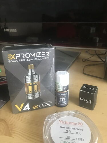 Sur la table, la boîte d'un atomiseur EXPromizer V4, une bouteille d'arôme STR, une boîte de pièces de rechange EXvape et du fil de résistance Nichrome 80 sont visibles. (Légendé par IA)