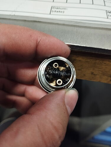 Egy kéz egy kis, kerek, elhasználódott vagy korrodált elektromos vagy mechanikus alkatrészt tart, amelyen két érintkező látható. (Feliratozva AI-val)