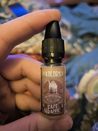 A képen egy "Kolors" márkájú, "Café Frappé" feliratú, kis üveg cseppentő látható, amelyet egy kéz tart. (Feliratozva AI-val)