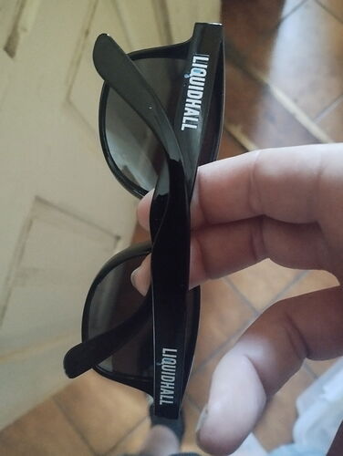 A képen egy kézzel tartott, fekete LIQUIDNALL feliratú napszemüveg látható. (Feliratozva AI-val)