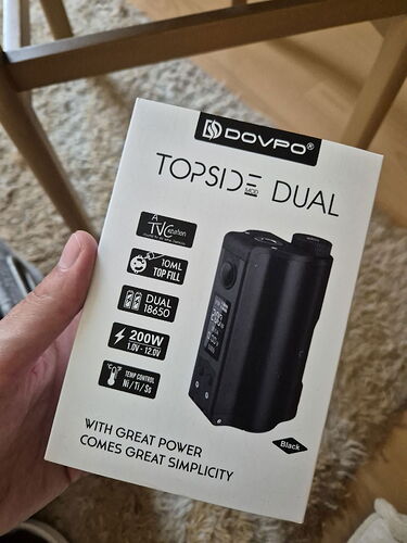 Une main tient une box de vaporisateur DOVPO TOPSIDE DUAL à puissance réglable électroniquement, d'une puissance de 200W et d'une capacité de remplissage de 10 ml. (Légendé par IA)