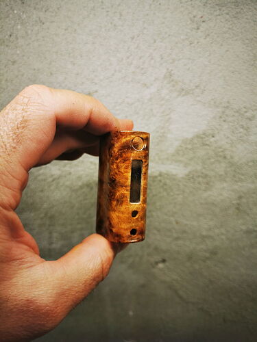 Une main tenant un appareil électronique en forme de boîte à motif de bois devant un fond gris. (Légendé par IA)