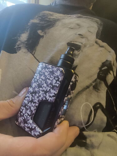 Une main tient une cigarette électronique à motifs noir et blanc, et en arrière-plan on voit un t-shirt noir et blanc sur lequel se trouve le portrait d'un chanteur à côté d'un microphone. (Sous-titré par IA)